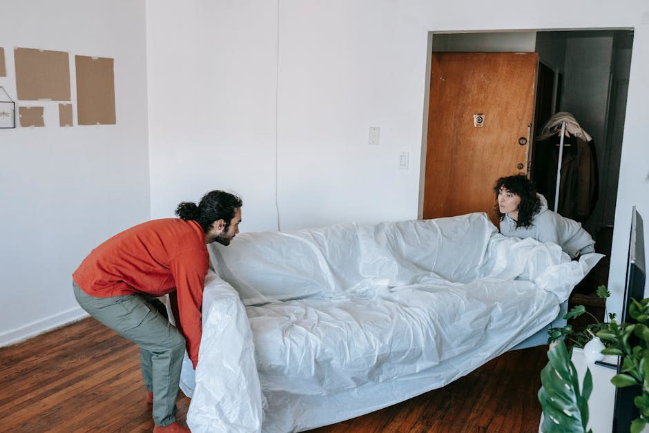 A man with dark, curly hair, wearing a red long-sleeved shirt and light-colored trousers, is carefully lifting and carrying a white sofa wrapped in protective packing material through a doorway inside a home. Inside the room, a woman with curly hair, dressed in a light-colored top, is reclining on the same sofa, observing the process. The room features white walls, a wooden floor, and a partially visible wall with some white adhesive notices. Adjacent to the doorway is a coat rack with a coat hanging on it. In the foreground, there is a green houseplant on a piece of furniture, and in the background, a wooden door is slightly ajar. The scene captures an active furniture transport or home relocation process, consistent with professional removals by Man with Van Brent Park.