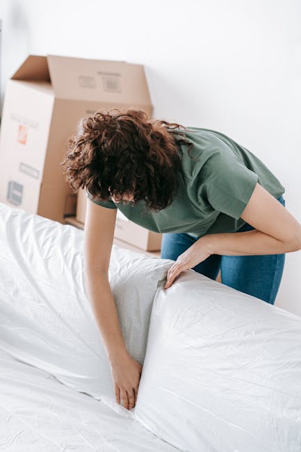A man with dark, curly hair, wearing a red long-sleeved shirt and light-colored trousers, is carefully lifting and carrying a white sofa wrapped in protective packing material through a doorway inside a home. Inside the room, a woman with curly hair, dressed in a light-colored top, is reclining on the same sofa, observing the process. The room features white walls, a wooden floor, and a partially visible wall with some white adhesive notices. Adjacent to the doorway is a coat rack with a coat hanging on it. In the foreground, there is a green houseplant on a piece of furniture, and in the background, a wooden door is slightly ajar. The scene captures an active furniture transport or home relocation process, consistent with professional removals by Man with Van Brent Park.