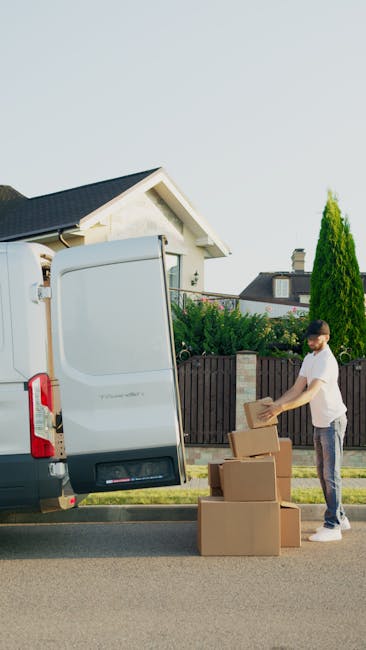 A man in casual attire standing beside a white moving van with its rear door open, loaded with several cardboard boxes of various sizes stacked on the paved street outside a residential property. The 