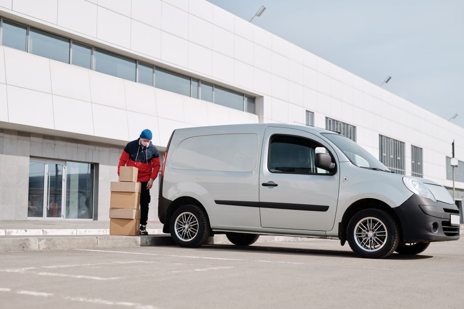 A man in casual attire standing beside a white moving van with its rear door open, loaded with several cardboard boxes of various sizes stacked on the paved street outside a residential property. The 