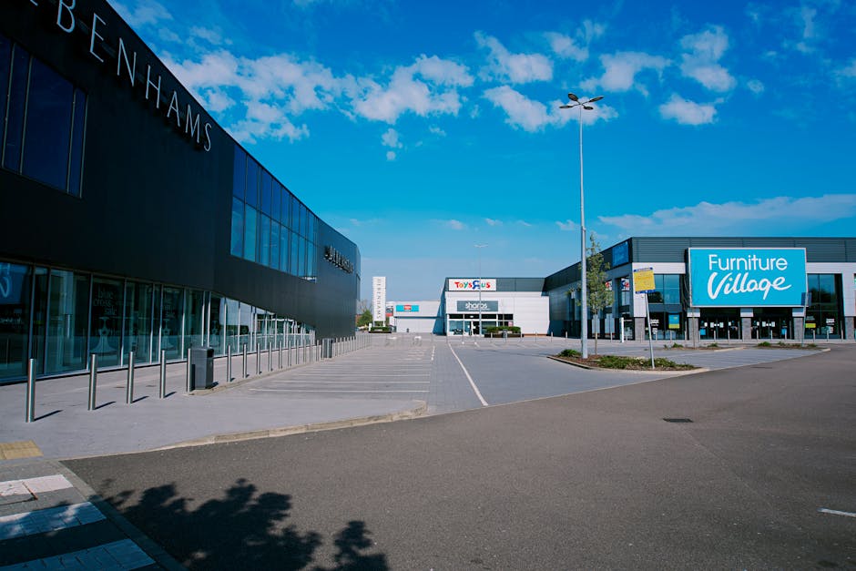 An empty car parking area outside retail units situated in Brent Park, featuring a large modern building with a dark exterior on the left displaying the name 'Benham's,' and another building on the right with a bright blue sign reading 'Furniture Village.' The scene is captured during daylight with a clear blue sky and scattered clouds overhead. The parking lot is largely vacant, with only a few small signs and streetlights visible, and the pavement is marked with white lines for parking spaces. This image depicts a typical commercial area where home relocation and furniture transport services, such as those offered by Man with Van Brent Park, may assist with loading and moving belongings during a house or retail unit removal process involving loading bay tips as outlined in the 'Moving out of Brent Park Retail Park' guide.