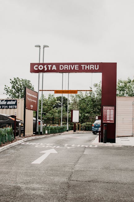 The image shows the entrance to a Costa Drive Thru located at the boundary of a retail park, with a large, red rectangular archway signage overhead displaying the Costa logo and the words 'DRIVE THRU.' Beneath the arch, a white arrow painted on the tarmac directs vehicles to proceed forward through the entrance. To the left side of the entrance, there are several small green park benches and a black car parked nearby. On the right, a white temporary building or storage unit is visible, along with a dark blue car waiting in the driveway. The scene is set during daytime with overcast skies, and the area is surrounded by greenery, including trees behind the infrastructure. The photograph is related to house removals or moving services, illustrating a typical vehicle access point at a retail park, which [COMPANY_NAME], based at [DOMAIN], may utilize for client relocations or furniture transport from commercial areas like Brent Park Retail Park, as discussed on the page titled 'Moving out of Brent Park Retail Park: loading bay tips, BRENT PARK' available at [PAGE_URL].