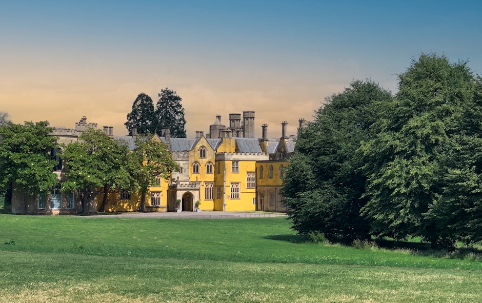 A large, historic yellow manor house with multiple turrets, chimney stacks, and detailed architectural features, situated behind a lush green lawn and trees. The property is surrounded by mature deciduous trees, with some in full leaf and providing partial shade, indicating a spring or summer setting. The scene is captured during late afternoon or early evening, with the sky showing a gradient from light to darker blue. The area appears peaceful and well-maintained, suitable for a high-end residence or estate. The image relates to house removals or home relocation services provided by Man with Van Brent Park, illustrating the type of property that may require furniture transport and packing during a move to Claremont Estate, Brent Park, as part of the local removals guide on their website.