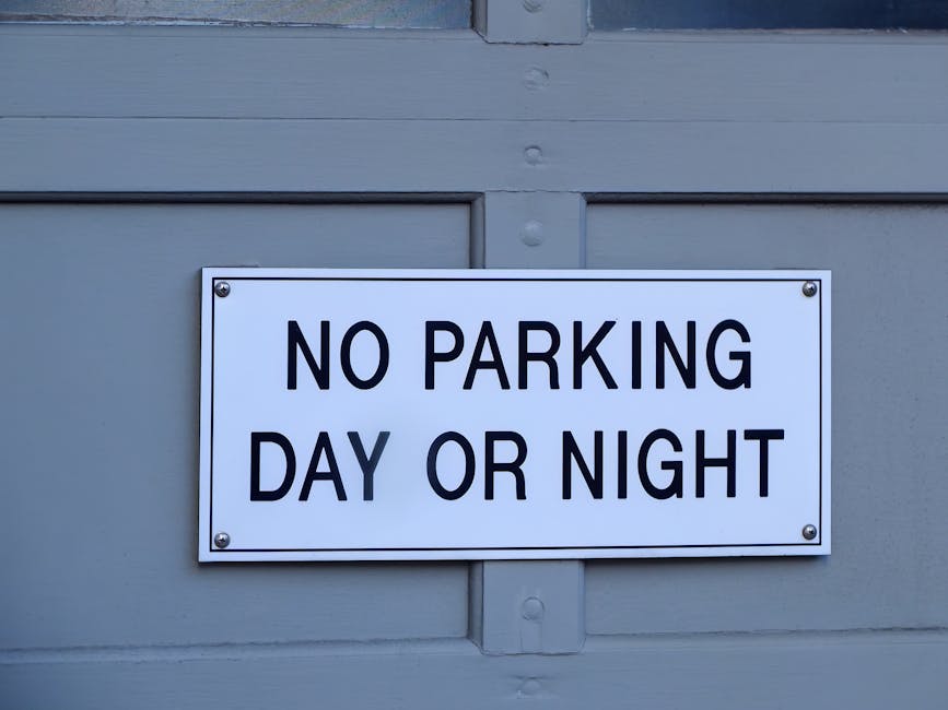 A white rectangular parking restriction sign with black bold lettering that reads 'NO PARKING DAY OR NIGHT' is mounted on a grey wooden wall, likely part of a garage or building exterior. The sign is secured with four visible screws, one in each corner. The background shows horizontal wooden panels painted in a light grey color, with some visible joints and textured wood grain. The sign's message indicates parking limitations in the area, which could be relevant for home relocation or furniture transport activities involving Man with Van Brent Park, especially under NW10 parking suspension rules for Brent Park movers. The scene is evenly lit, highlighting the clarity of the sign and the surrounding environment, emphasizing the importance of adhering to local parking regulations during moving or packing processes.