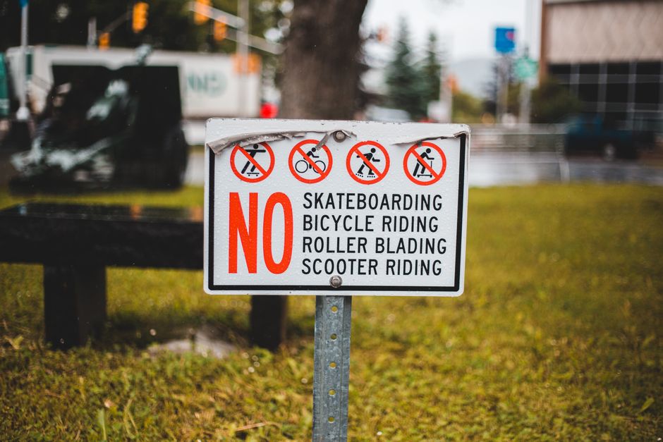 A rectangular outdoor sign mounted on a metal post in a grassy area with a paved roadway and trees in the background. The sign features four red circle icons with diagonal lines across illustrations indicating no skateboarding, no bicycle riding, no roller blading, and no scooter riding. Below these icons, the text reads “NO” in large red capital letters, followed by black text listing the prohibited activities: ‘SKATEBOARDING,’ ‘BICYCLE RIDING,’ ‘ROLLER BLADING,’ and ‘SCOOTER RIDING.’ The lighting appears natural, and the surrounding environment includes a black vehicle and blurred background of buildings and traffic lights. This sign is relevant to legal restrictions in residential or street areas, often considered during house removals or loadings when parking or access is limited by local rules, as seen in the context of house moves. Man with Van Brent Park ensures adherence to local regulations during furniture transport or packing and moving activities, as illustrated in the image.