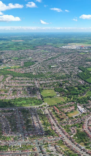 An aerial view of a residential area showing numerous houses arranged along tree-lined streets, with some properties featuring garages and driveways. There are patches of green spaces, sports fields, and small parks scattered throughout the suburb. The scene captures the edge of the urban environment with a mix of built-up areas and open spaces, under a bright blue sky with a few scattered clouds. In the foreground, part of a street is visible, and within the context of house removals and relocation services, this image illustrates the typical environment where furniture transport, packing, and loading processes by Man with Van Brent Park might take place during a home relocation or moving operation.