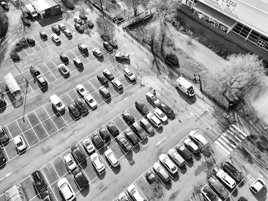 An aerial black-and-white photograph capturing a large parking lot filled with numerous parked cars arranged in neat rows, with visible white lines marking individual parking spaces. A few empty spaces are dispersed throughout the lot. Surrounding the parking area, there are trees casting shadows on the pavement. On one side, there is a building with large windows and a street with a pedestrian crossing, where a white van from Man with Van Brent Park is parked near an entrance, likely engaged in a home relocation or furniture transport process. The van is positioned close to a group of packing materials, possibly cardboard boxes and furniture blankets, indicating the ongoing loading or unloading activity typical of house removals. The overall scene suggests a busy, well-organized environment focused on efficient moving logistics, with clear evidence of packing, parking, and transportation elements within a typical urban setting.