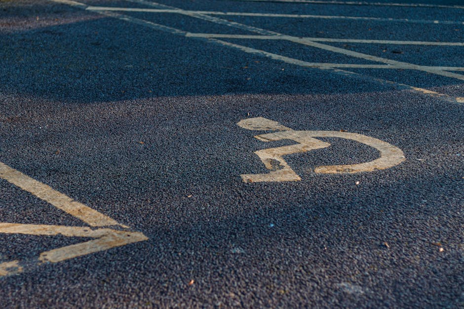 A black-and-white aerial photograph of a large parking lot adjacent to a building, with multiple evenly spaced parking bays and white painted lines designating individual parking spaces. Several cars are parked throughout the lot, with some in designated bays and others in areas reserved for loading or disabled parking, indicated by specific markings on the asphalt. A few vehicles are parked close to the building entrance, which is not visible in the image. The parking lot is bordered by a row of trees on one side, providing natural separation from neighboring areas. The scene appears to be well-lit, with visible shadows suggesting daytime. This setting could be used for home relocation purposes where vehicles from Man with Van Brent Park load furniture and boxes for moving services, supporting efficient and organized transport during the packing and moving process.